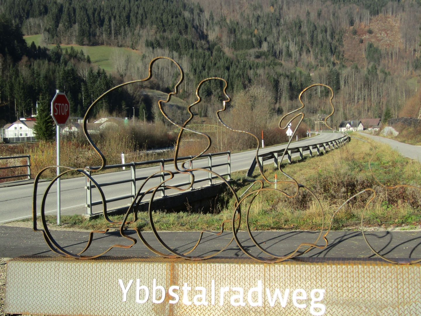 Silhouette von Radfahrern als Metallskulptur am Straßenrand des Ybbstal-Radwegs mit Stoppschild und Landschaft.