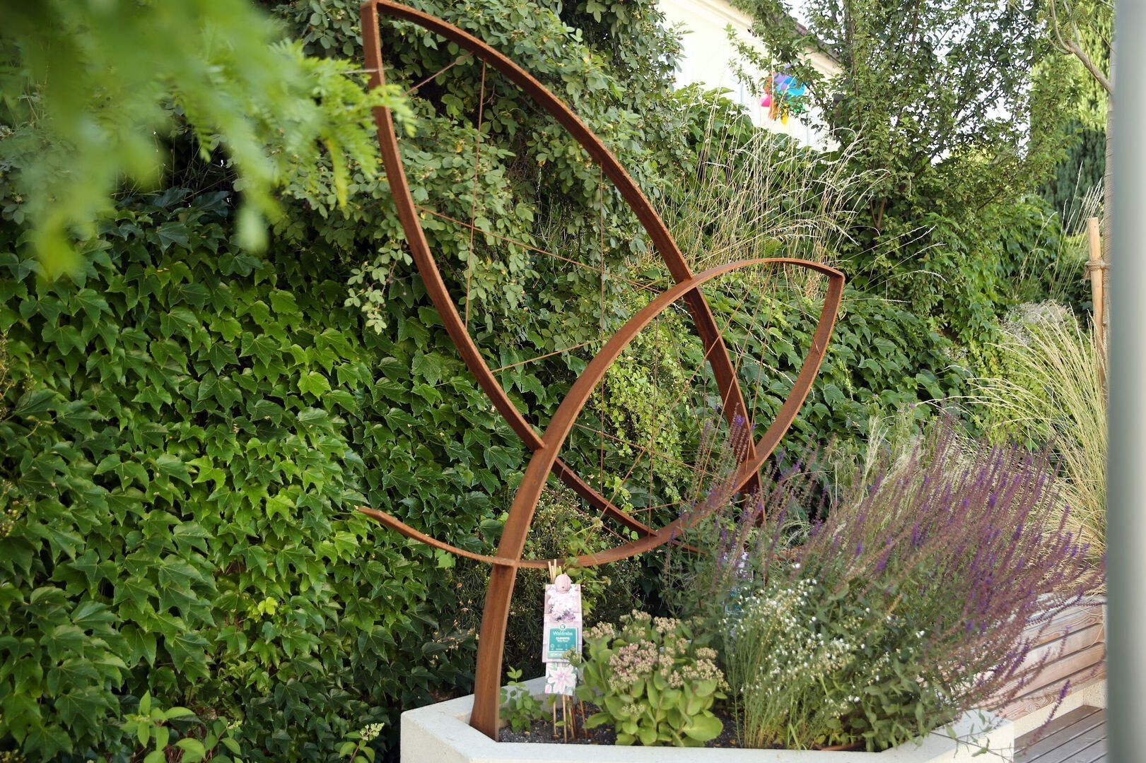 Kunstwerk aus Eisen in einem Pflanzkübel aus Beton vor einer efeubewachsenen Wand