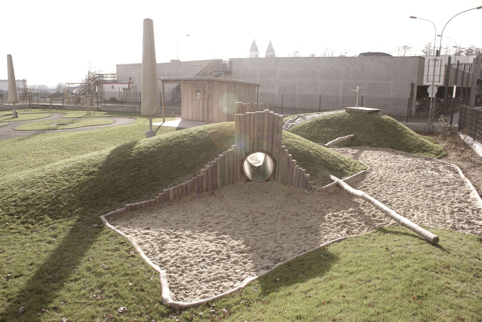 Spielplatz mit begrasten Hügeln, Sandkasten, Holzpalisaden und hölzernen Balancierbalken