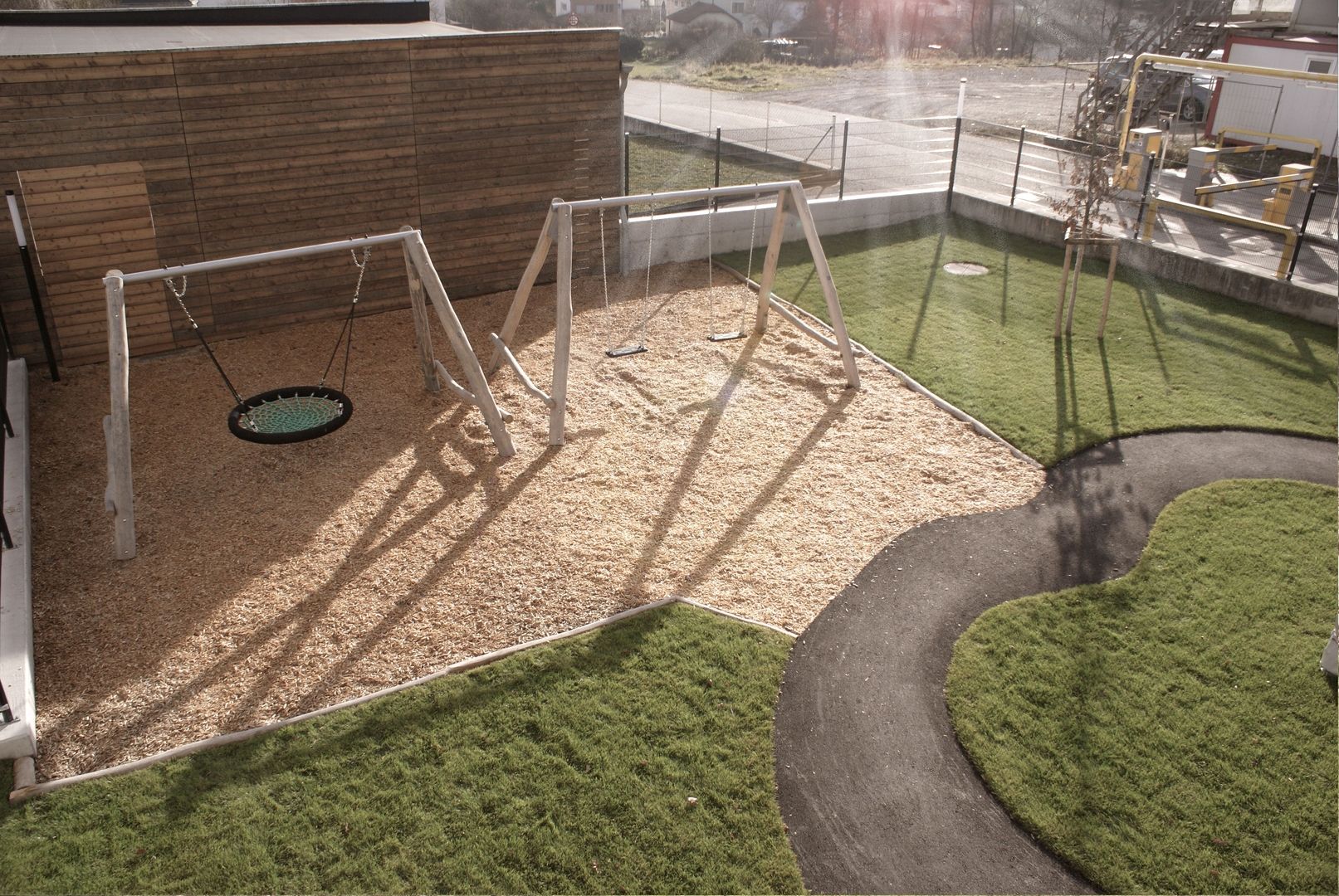 Kinderschaukeln an Holzgerüsten in einem von einer Wiese umrahmten Sandkasten vor einer Holzhütte