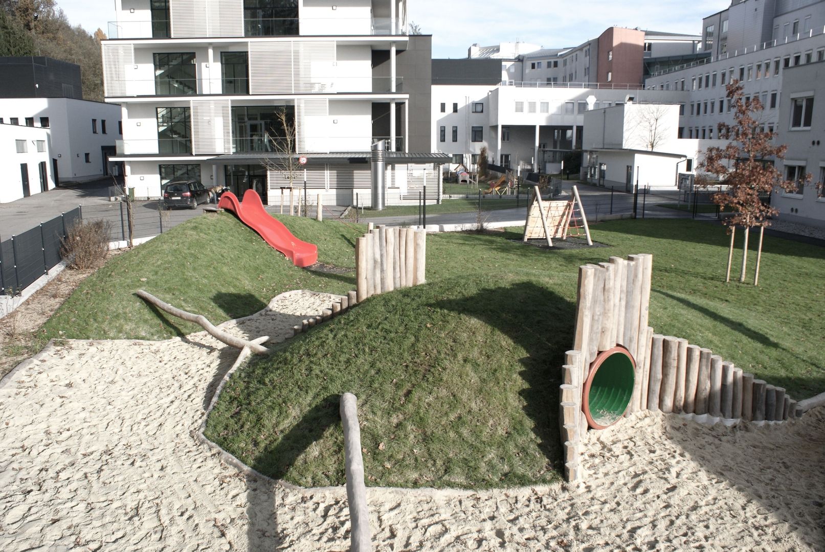 Spielplatz mit begrasten Hügeln, Sandkasten, Holzpalisaden und hölzernen Balancierbalken in einem Wohngebiet