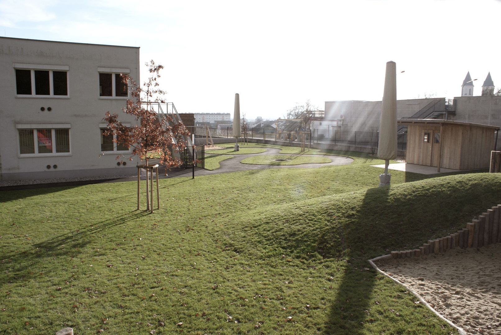 Sonniger Spielplatz mit Gras, Wegen, Bäumen und Gebäuden im Hintergrund.