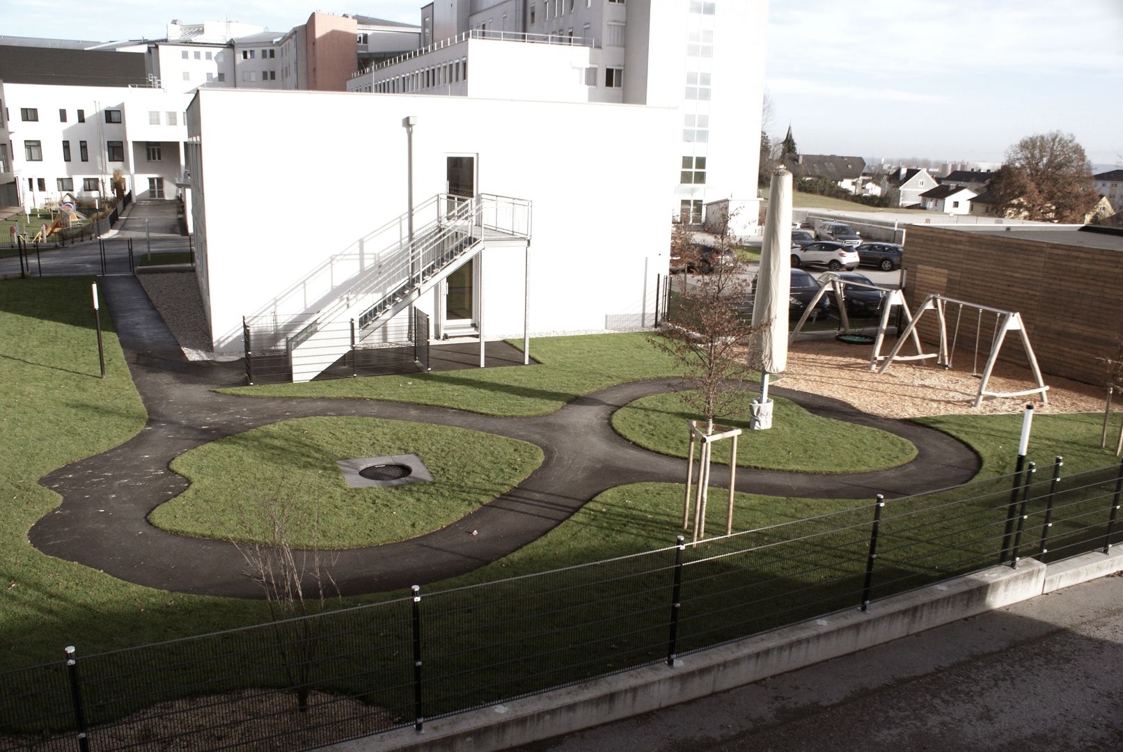 Spielplatz im Freien mit gewundenen Wegen, Schaukeln und Grünflächen, neben einem Gebäude