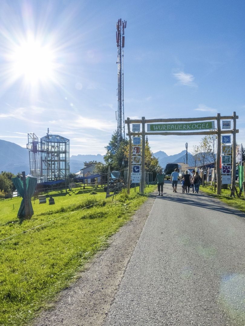 Sonniger Weg, der zum Eingang des Wurbauerkogels führt, mit Spaziergängern und einem Mobilfunkmast auf der linken Seite