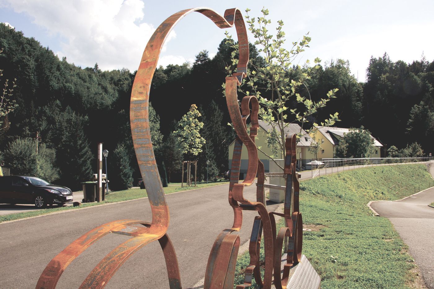 Silhouette von Radfahrern als Metallskulptur am Straßenrand auf einer Straße mit Bäumen und einem Auto im Hintergrund